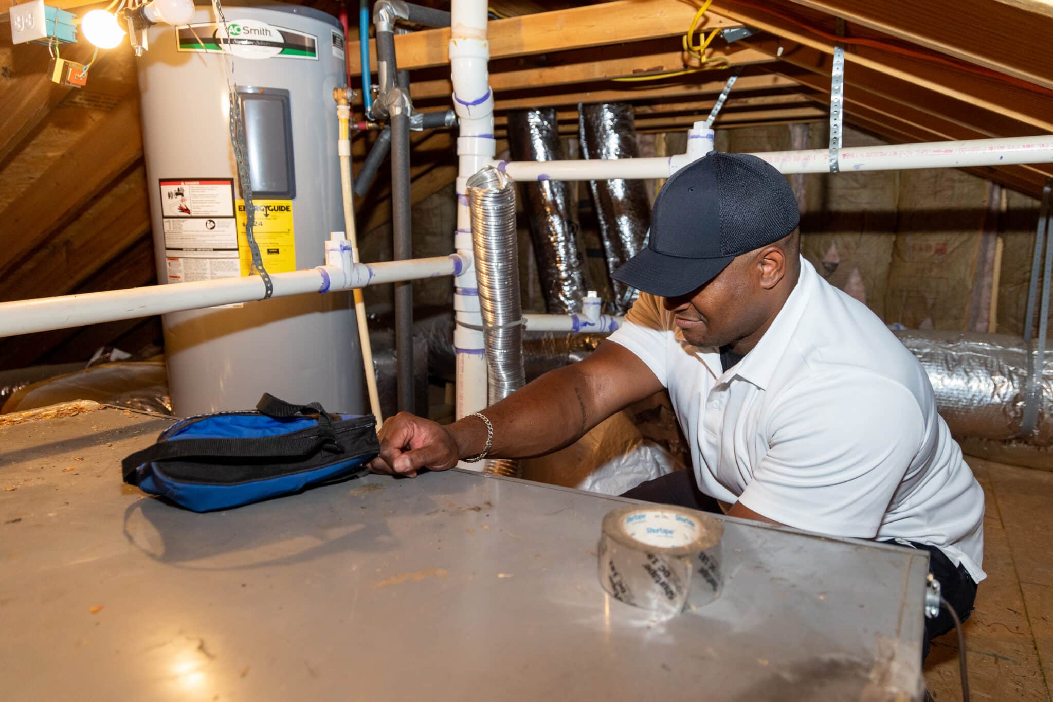 HVAC technician servicing a whole-home air purifier connected to ductwork in an attic.
